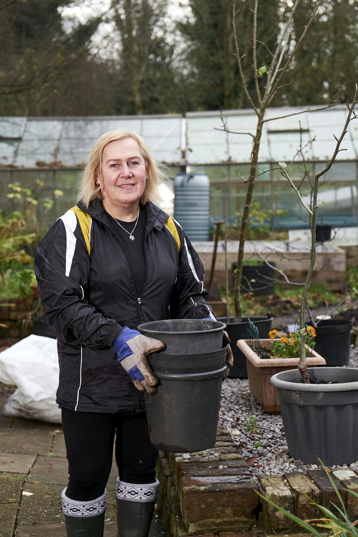 Woman gardener in a garden holding pots. Greenhouse and plants in the background.